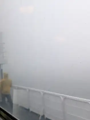 Through a misted, rain-spotted ferry window, a person in a yellow hooded jacket stands at the white deck railing facing a wall of fog.