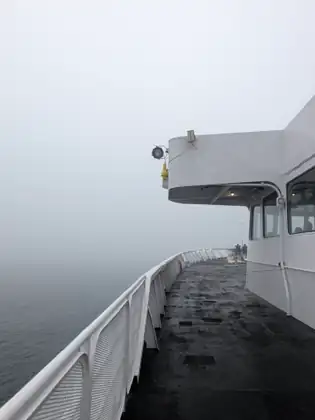 The ferry's wet outer deck runs along a white railing beneath an overhanging bridge wing, with two people at the far end and the sea fading into thick fog.