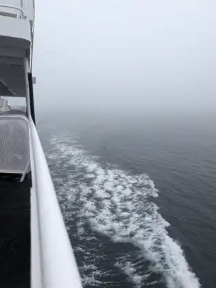White wake churns along the side of the ferry below a white railing as the grey sea disappears into fog.