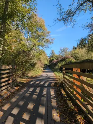 A wooden trail bridge with slatted rails throws striped shadows across its deck, leading on toward a paved path through autumn trees.