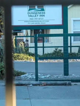 The Dungeness Valley Inn sign, giving its address as Carlsborg Road, tops a green-framed wire fence in front of a pale yellow building, seen past a blurred post in the foreground.