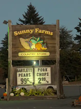 At dusk, the lit Sunny Farms Country Store sign, carved with a cornucopia of produce, stands above a board advertising Bartlett pears at 99 cents a pound and pork chops at $2.99 a pound, with pumpkins at its base.