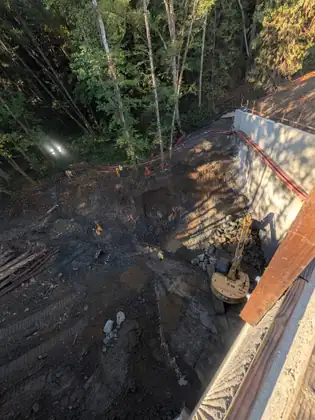 Looking straight down over a concrete edge into a deep construction pit, where an excavator sits beside a tall concrete wall and a pile of boulders, red hoses run along the rim, and workers in high-visibility gear stand near bright work lights at the edge of the woods.