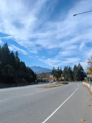 A highway splits under a blue sky streaked with wispy cloud, a lane curving right between a low concrete barrier and a guardrail while the main road bends left past road signs and distant traffic toward dark evergreens, autumn trees and a hazy mountain.