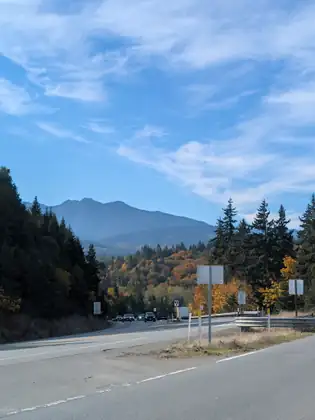A highway runs past road signs and autumn trees toward distant traffic, with a hazy blue mountain ridge rising behind under a blue sky.
