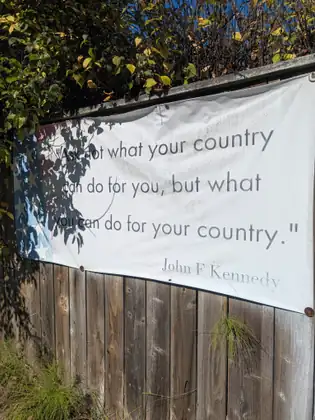 A white banner hung on a weathered wooden fence, dappled with leaf shadows and printed with a faded American flag at one end, quotes John F Kennedy: "Ask not what your country can do for you, but what you can do for your country."