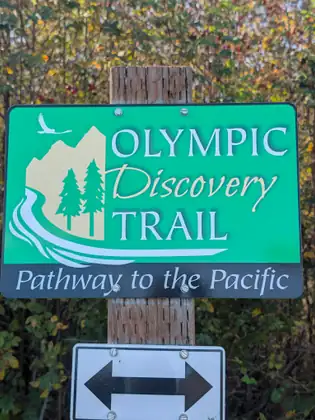 A green sign on a wooden post reading Olympic Discovery Trail, Pathway to the Pacific, with a mountain, two evergreen trees and a flying bird in its logo, above a double-headed arrow.
