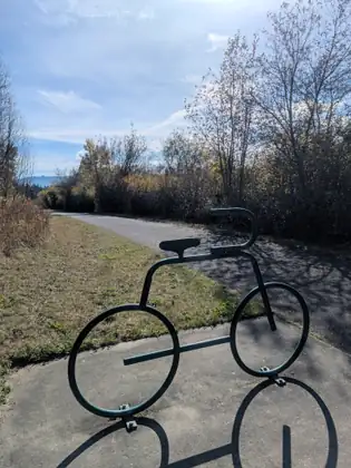A bike rack shaped like a bicycle outline in dark green metal tubing, bolted to concrete beside a paved trail that curves away past autumn shrubs under a blue sky.