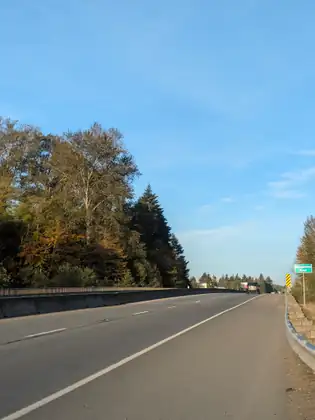 A highway running ahead under a blue sky, with a concrete barrier along its left side and tall autumn trees beyond, an SUV in the distance and a green sign reading Dungeness River by the guardrail on the right.