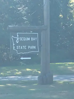 A dark brown wooden sign painted with a white outline of Washington State and the words Sequim Bay State Park hangs from a log arm on a tall wooden post, above a small sign with a white arrow pointing left, in hazy light.