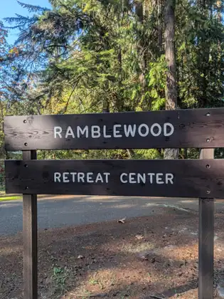 A dark wooden sign of two planks reading Ramblewood Retreat Center stands beside a paved driveway under tall evergreens and sunlit trees.