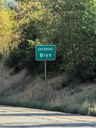 A green road sign reading Entering Blyn on a metal post at the edge of a highway, in front of a grassy bank and leafy trees.