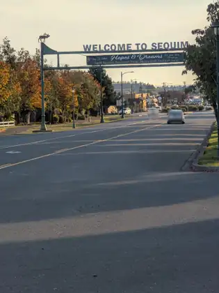 A gateway over a wide road reading WELCOME TO SEQUIM, an antlered animal cut-out on its left post and a banner reading Hopes & Dreams, Sequim Education Foundation's Annual Teaching Grant Fundraiser Dinner, Oct. 24th, 2026, with cars on the road beyond.