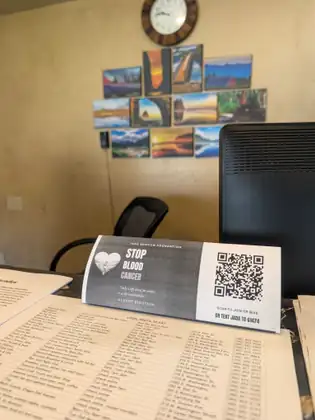 A Jada Bascom Foundation table tent reading STOP BLOOD CANCER, with a QR code and the words Scan to join or give or text JADA to 61474, standing on a front desk over a printed list headed Local Spots to Eat.