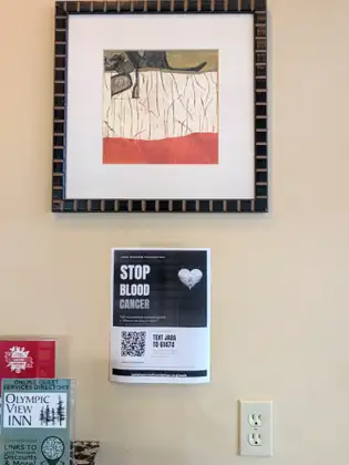 A Jada Bascom Foundation poster reading STOP BLOOD CANCER, TEXT JADA TO 61474 and jadabascomfoundation.org/walk, hung on a cream wall below a framed print, beside an Olympic View Inn guest services sign.