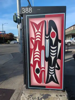 A street-corner utility cabinet numbered 388, painted with two stylised fish side by side, one red and one black, in bold red, black, white and pale blue shapes, with a traffic signal pole behind.