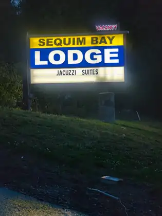 A lit roadside sign reading Sequim Bay Lodge, Jacuzzi Suites, with a red Vacancy sign glowing above it, stands on a grassy bank in the dark beside a wet road.