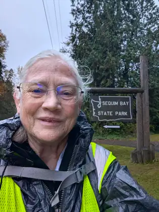 Jeana Moore, her white hair blown about, smiles in a black rain jacket and hi-vis vest with pack straps across her chest, in front of a wooden sign reading Sequim Bay State Park above an arrow pointing left.