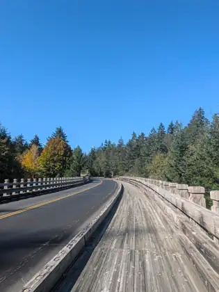 A bridge with weathered timber railings curves away under a cloudless blue sky, a wide plank walkway running beside its two traffic lanes and double yellow centre line toward dark conifers and yellow autumn trees.