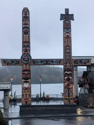 Two tall carved and painted poles, with a carved crossbeam spanning them, rise from a stone base lettered Jamestown S’Klallam Tribe, with a rain-wet plaza, lamp posts and a misty bay and wooded hills behind.