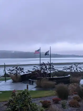 An American flag and a black flag fly from two poles above a curved dark wall lettered In Honor of Those Who Served and, partly hidden by grasses, Jamestown S’Klallam Veterans Memorial, with wet flats and misty wooded hills beyond.