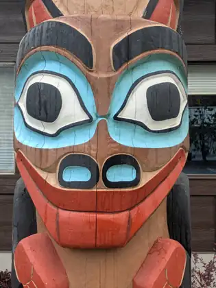 A close-up of a face carved into a wooden pole, with round black-and-white eyes ringed in turquoise, heavy black brows, turquoise nostrils and a wide red grin, wet with rain in front of a dark-sided building.