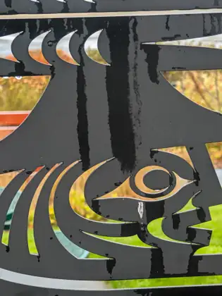 A close-up of a rain-wet black metal panel cut into a stylised design of an eye and curving crescents, with orange shrubs, red slats and green lawn showing through the openings.