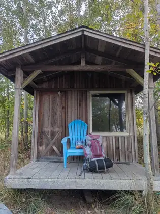 A small weathered wooden cabin with a peaked roof and covered porch, where a bright blue Adirondack chair holding a clear bag of fruit stands beside a packed maroon and gray backpack marked STEPS TO MARROW, among slender trees.