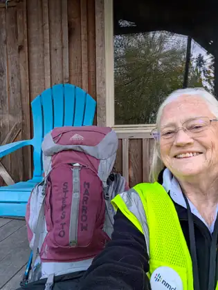 Jeana Moore in a hi-vis vest smiles into the camera on a wooden porch beside her maroon and gray backpack, the words STEPS TO MARROW written down its front, in front of a bright blue Adirondack chair.
