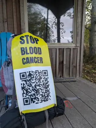 A yellow and white STOP BLOOD CANCER banner with a large QR code hangs from the back of a packed backpack on a wooden porch, a blue chair and a cabin window behind it.