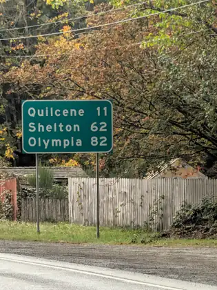 A green highway sign reading Quilcene 11, Shelton 62, Olympia 82 stands on the grassy roadside in front of a weathered wooden fence and trees turning orange and gold.