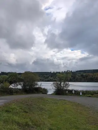 Heavy gray and white clouds fill the sky over a stretch of water ringed by forested hills, with a gravel lot and grassy bank in front and three small signs by the water, one reading Discover Pass Required.