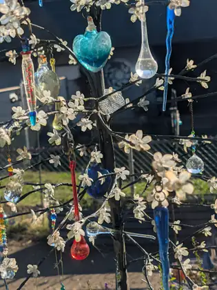 A decorative metal tree covered in small white artificial blossoms and hung with glass ornaments, among them a turquoise heart, clear teardrops, blue icicles, a red drop, a cobalt heart and cut-crystal prisms, glinting in the sun before a chain-link fence.