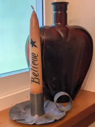 A tan taper candle hand-lettered Believe beneath a small black star stands in a tin chamberstick with a ring handle, in front of a heart-shaped amber glass bottle on a wooden windowsill.