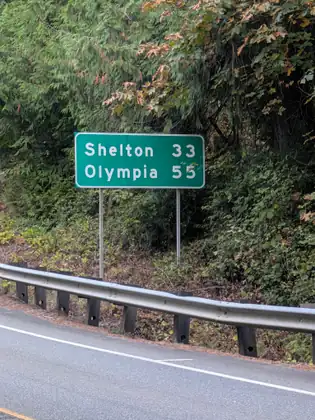 A green highway sign reading “Shelton 33, Olympia 55” stands behind a metal guardrail on a bend of the road, against a steep bank of ferns, maples and evergreens.