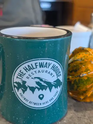 A speckled teal enamel mug printed with the Halfway House Restaurant logo and the words “Halfway between here and there” sits on a table beside a small orange-and-green striped gourd.
