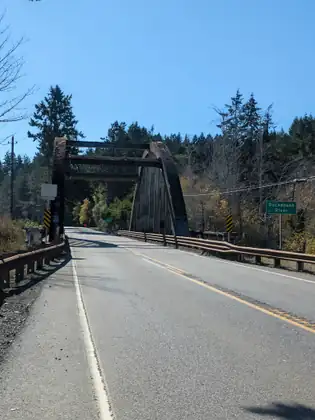 Highway 101 runs onto an old steel truss bridge, with a green “Duckabush River” sign on the right, guardrails along both sides and tall evergreens beyond under a clear blue sky.