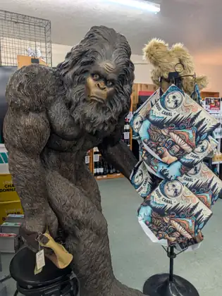Inside a shop, a life-size bronze-coloured Bigfoot statue stands beside a rack of oven mitts printed with a blue mountain pattern.