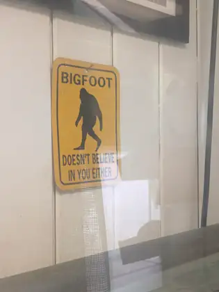 A yellow sign in a window shows a walking Bigfoot silhouette and reads “Bigfoot doesn’t believe in you either.”