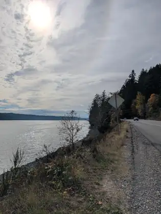 The highway runs along the edge of the bay under a bright, hazy sun, with a dry grassy bank, a road sign and wooded hills along the shore.