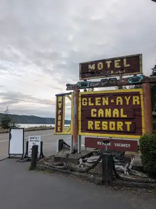 A carved wooden sign for the Glen-Ayr Canal Resort, topped by a Motel sign, with an RV Park sign and a vacancy board, stands beside the highway with the water behind.