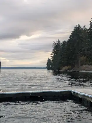 The corner of a wooden dock juts into calm water toward a dark wooded point under a pale evening sky.