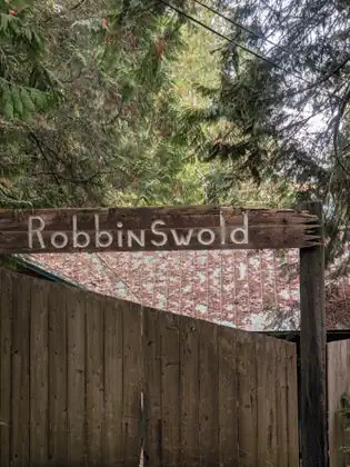 A weathered wooden sign carved with the name Robbinswold hangs above a tall wooden fence and gate among evergreen branches.