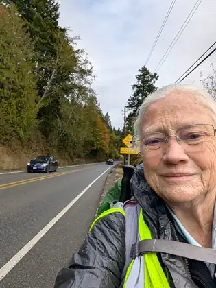 Jeana, smiling, with short white hair, glasses and a yellow-green safety vest over her jacket and pack straps, takes a selfie on the shoulder of a highway lined with autumn trees as cars pass.