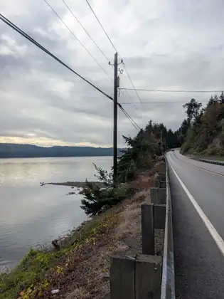 The highway runs along a wooden guardrail beside a calm bay, with power lines overhead and wooded hills across the water under a cloudy sky.
