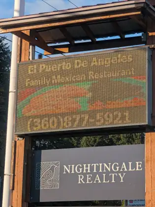 A roadside sign with a scrolling display for El Puerto De Angeles Family Mexican Restaurant and its phone number stands above a sign for Nightingale Realty.