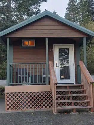 A small brown cabin with green trim, a little porch with chairs and wooden steps sits on gravel among trees.
