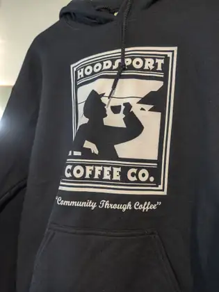 A black hoodie printed with the Hoodsport Coffee Co. logo, a figure sipping from a cup, and the words “Community Through Coffee.”