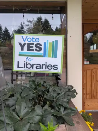 A green-and-white “Vote YES for Libraries” sign hangs in a shop window above a planter of dark green leaves.