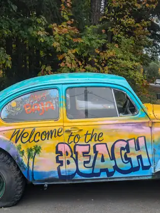 A vintage Volkswagen Beetle painted in sunset colours with palm trees and the words “Welcome to the Beach” is parked beneath autumn trees.